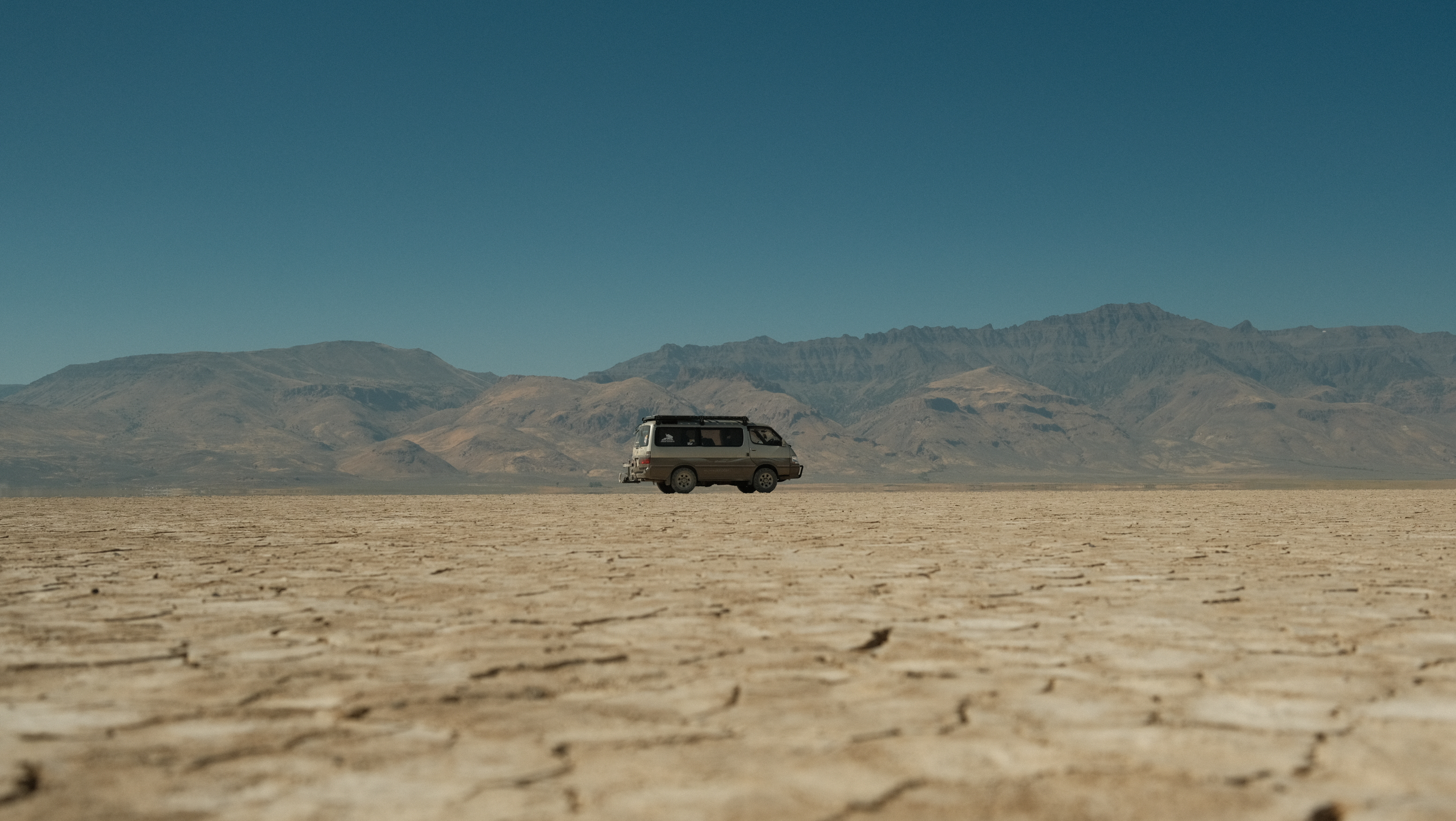 A tan mini-van sits placidly mid-frame. Beneath it is a cracked, tan desert playa. Behind it a mountain blocks the horizon. Above it is dusty blue skies. 